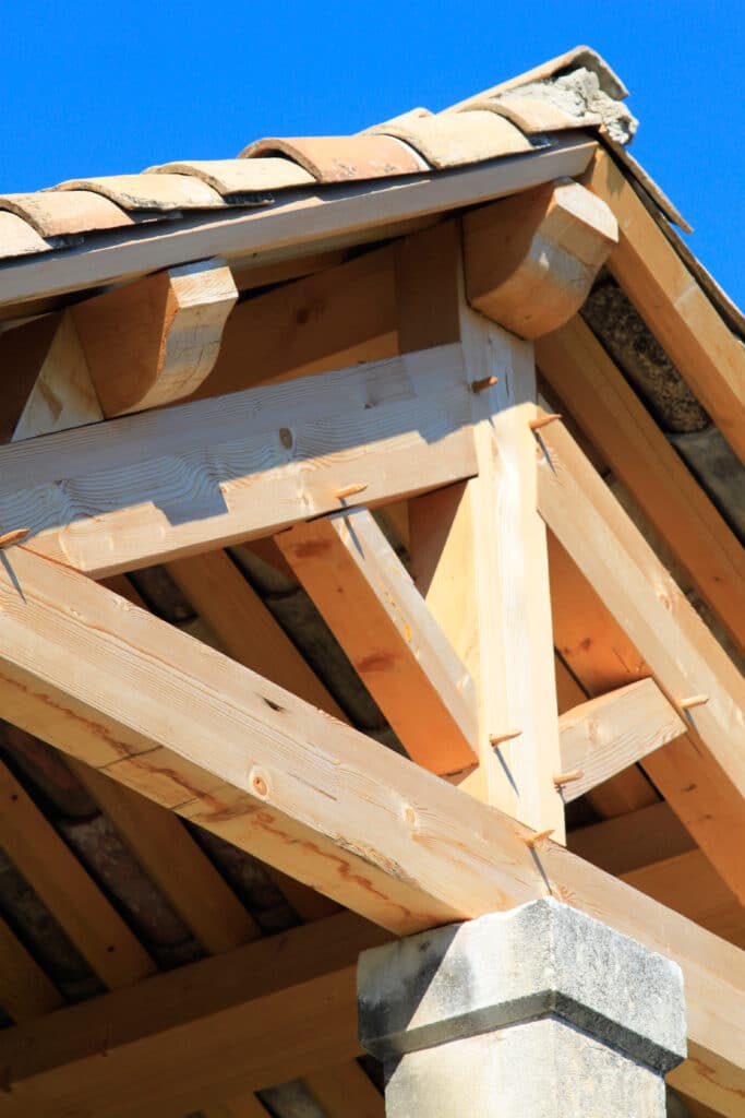 Charpente en bois et toit tuiles: Artisanat sous ciel bleu. Charpente artisanale en bois apparent avec chevilles, supportant des tuiles en terre cuite. Vue détaillée sous ciel bleu, sur pilier pierre.
