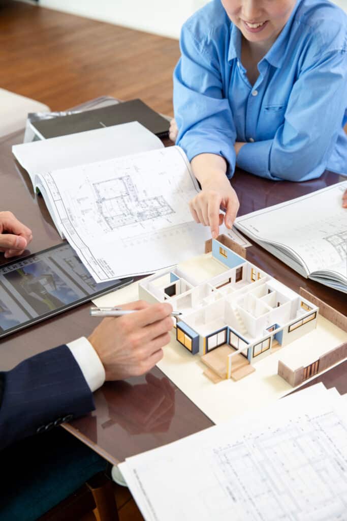Architecture maison: consultation plans et maquette 3D Consultation architecture: deux personnes (femme souriante, homme) examinent plans, maquette de maison et rendu 3D sur tablette. Ils pointent des détails.