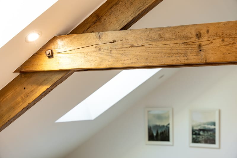 Design mansarde : Poutres bois rustiques, lumière naturelle. Poutres rustiques en bois dans une mansarde lumineuse, avec un spot encastré, une lucarne et deux tableaux flous en fond.